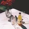 A vase of red roses on a table with a bottle of bittersweet liqueur and two empty glasses, set on a white tablecloth in sunlight.