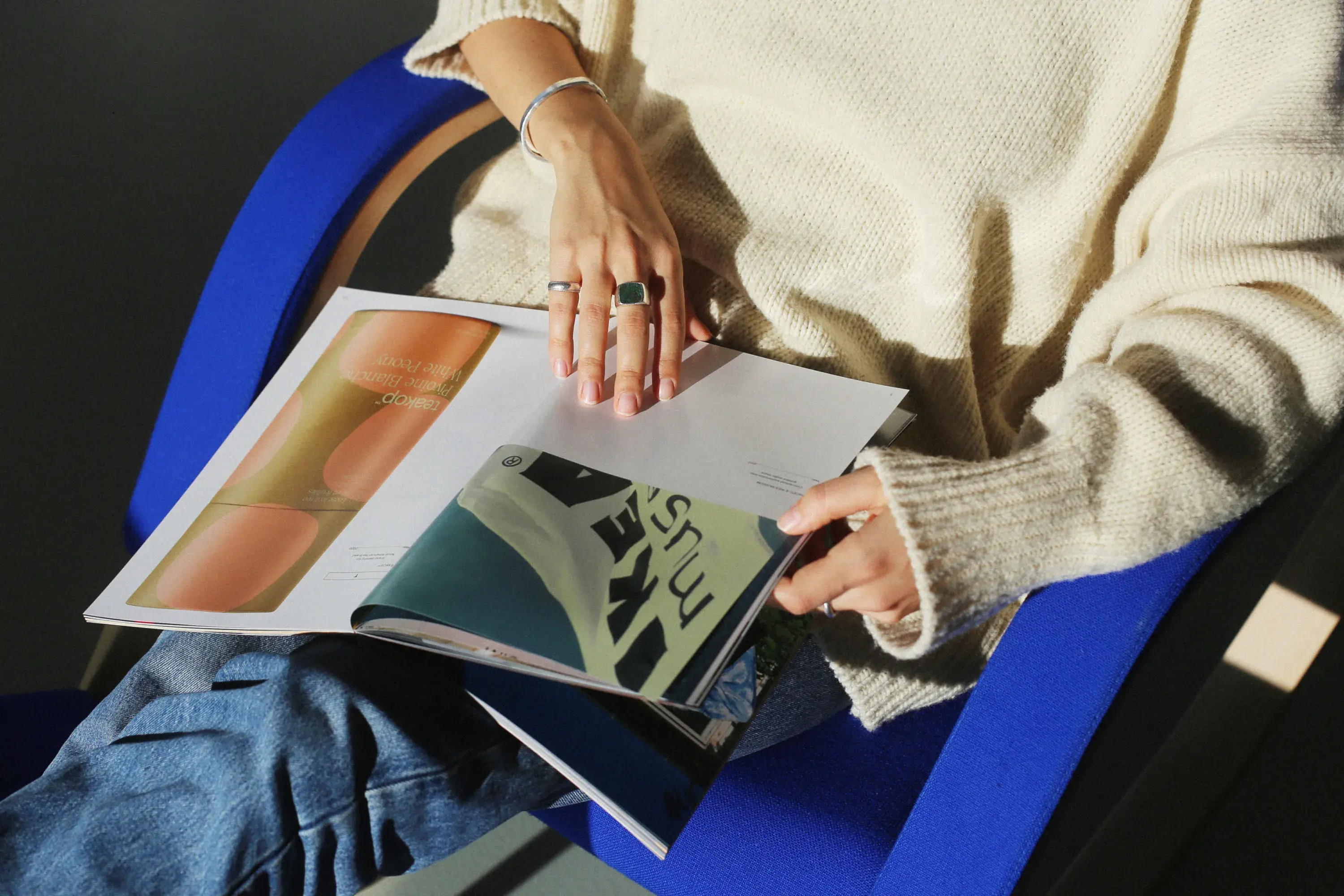 Person in a cream sweater and jeans sits on a blue chair, flipping through a magazine with colorful pages.