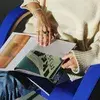 Person in a cream sweater and jeans sits on a blue chair, flipping through a magazine with colorful pages.
