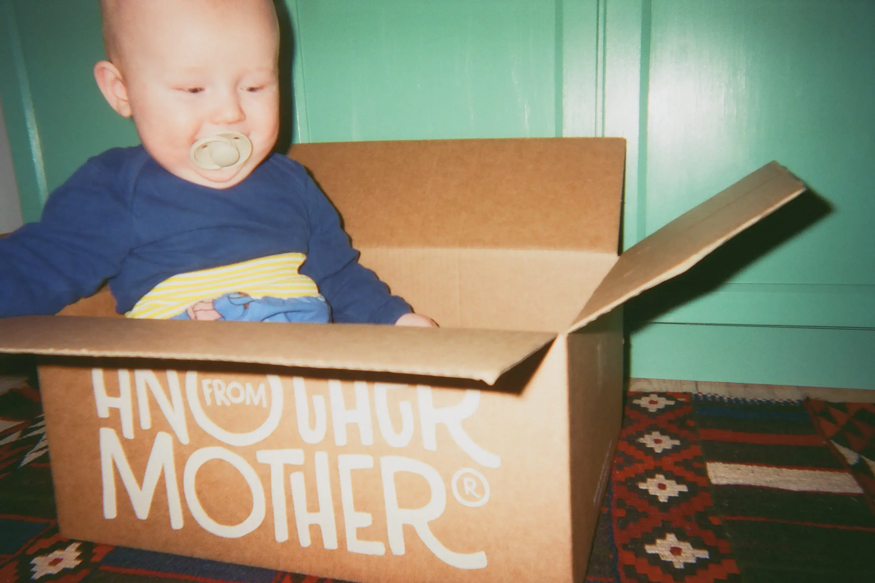 A baby with a pacifier sits in an open cardboard box on a patterned rug, in front of a green wall.