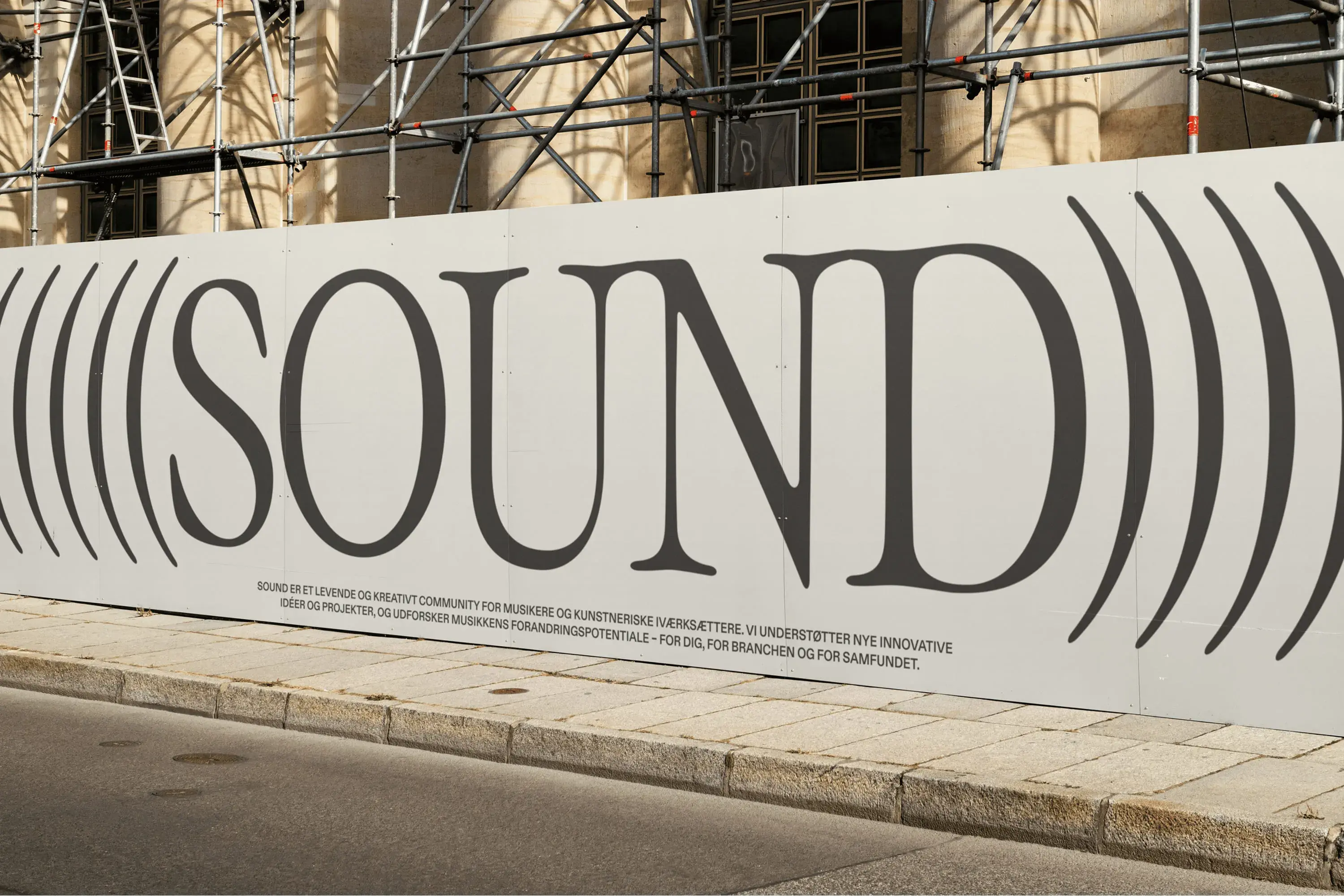 Large white sign with the word "SOUND" in bold letters, surrounded by sound wave graphics, placed on a street with scaffolding behind.